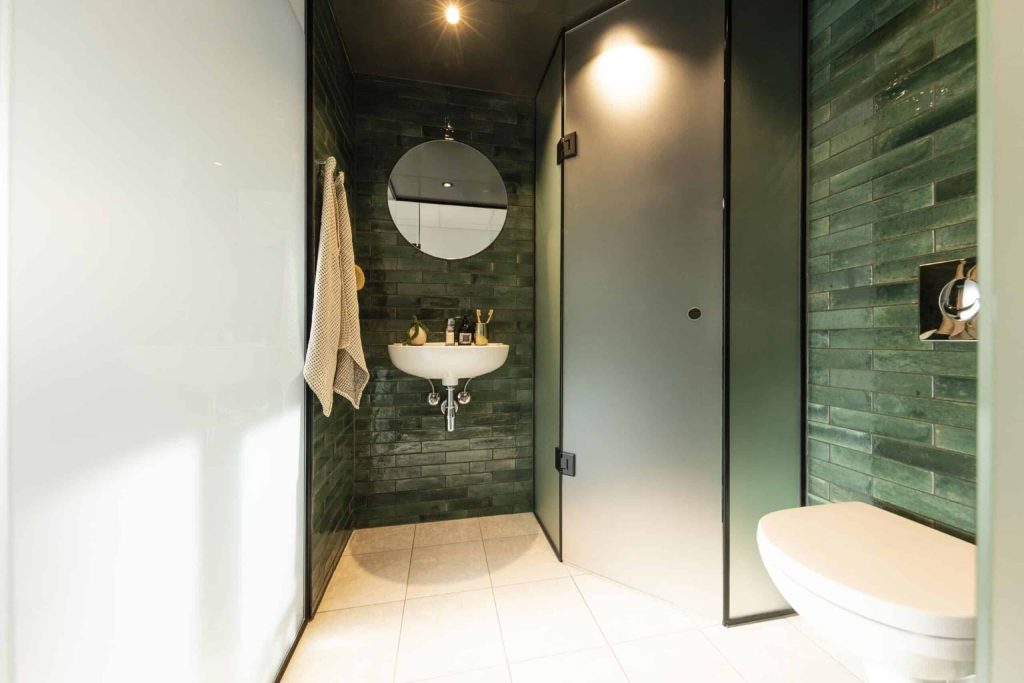 Moderne og stilrent Campus Aviator badeværelse med grønne fliser, rund spejl, væghængt håndvask, glasafskærmning og elegant belysning i lyst interiør.