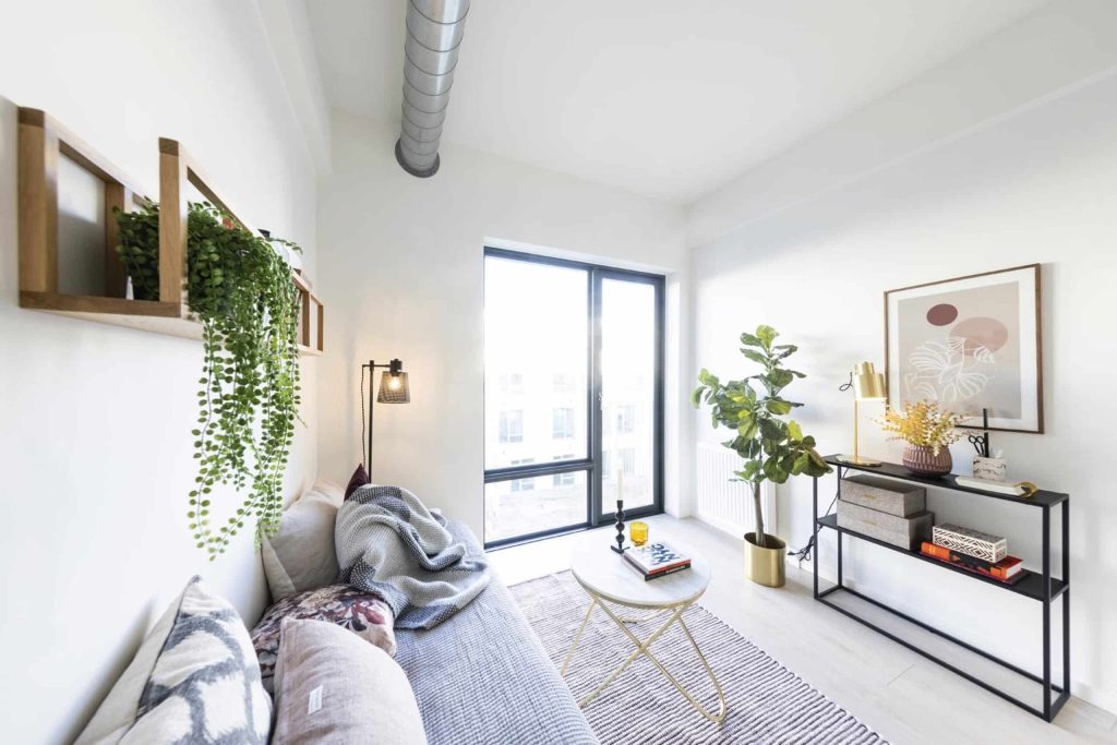 Lys stue med Campus Aviator udsigt gennem store vinduer, moderne indretning med sofa, planter og stilrent interiør, der giver masser af dagslys og åben rumfornemmelse.