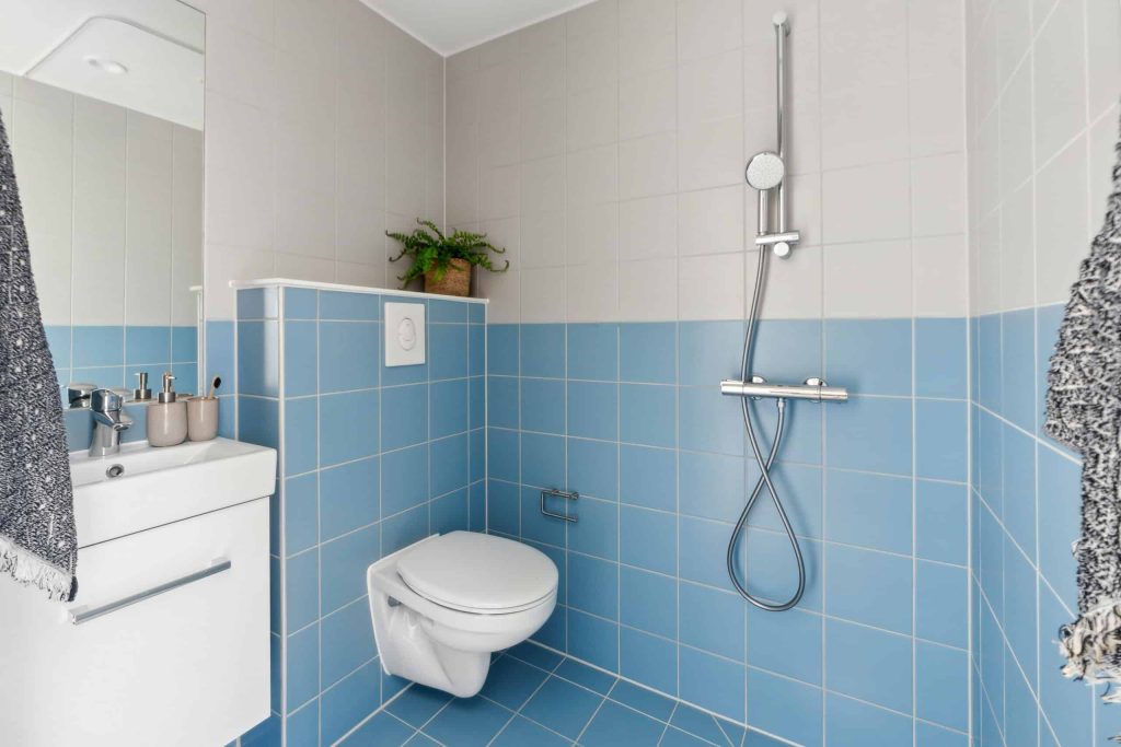 Lyst og funktionelt midgaard badeværelse med blå og lyse fliser, væghængt toilet, moderne brusearmatur, håndvask og minimalistisk indretning.