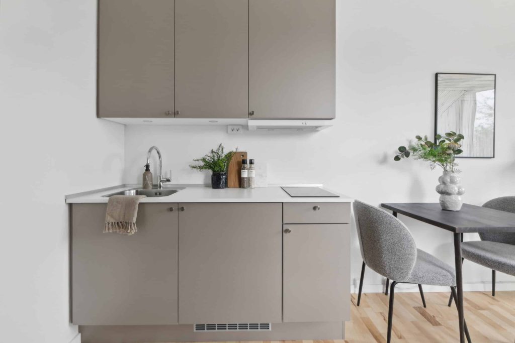 Stilrent og funktionelt midgaard køkken med grå skabe, integreret kogeplade, hvid bordplade, moderne armatur og spiseplads i lyst, minimalistisk interiør.