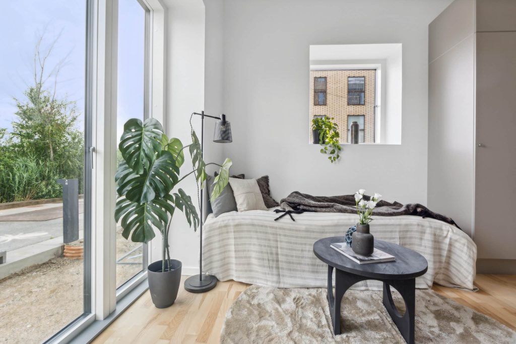 Lyst og roligt midgaard soveværelse med komfortabel seng, store vinduer, grønne planter, moderne belysning og stilren indretning med masser af dagslys.