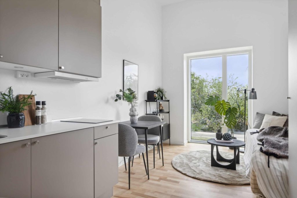 Lys og indbydende midgaard stue med åbent køkken-alrum, spiseplads, moderne indretning, grønne planter og store vinduer med udsigt til natur.